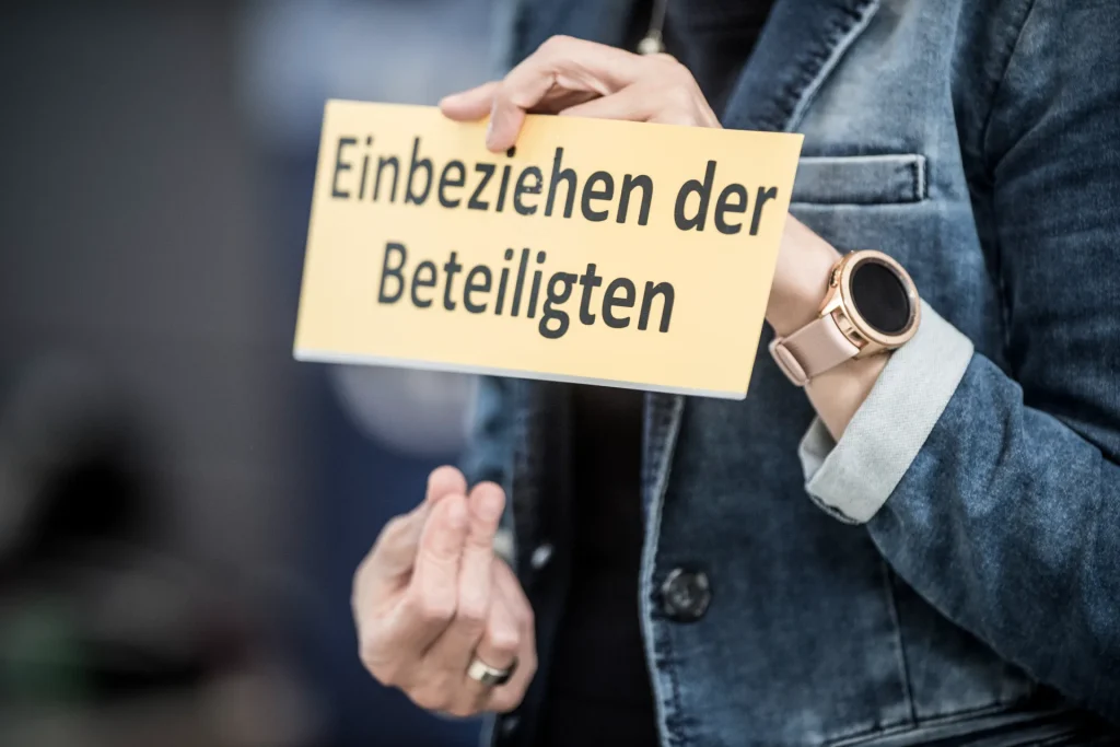 Hände halten Schild mit der Aufschrift "Einbeziehen der Beteiligsten" im gemba.coach Training